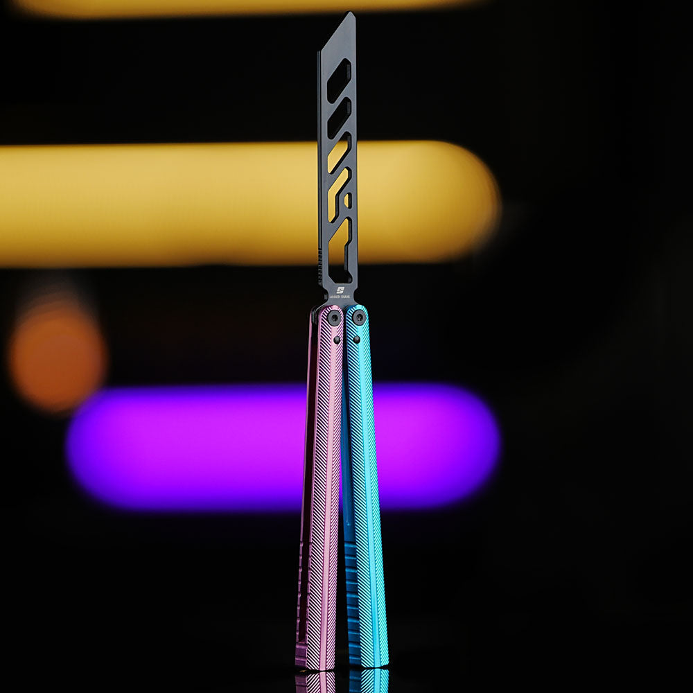 Armed Shark Pacific Ocean Balisong Trainer featuring pink-blue gradient anodized aluminum handles and black training edge, standing upright on reflective surface with vivid yellow and purple background lights