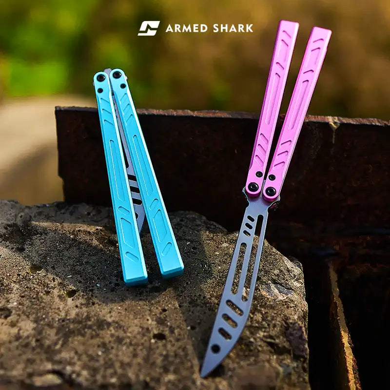 Armed Shark BB 7075 balisong trainers in blue and pink handles displayed outdoors