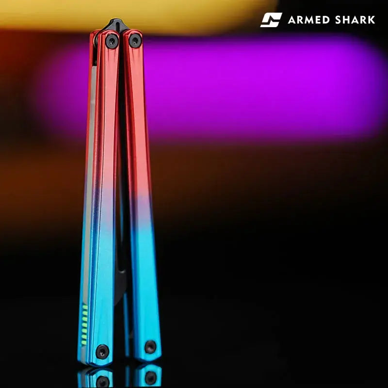 Folded view of Armed Shark Eureka Balisong Trainer in red-blue gradient anodized finish, showcasing precision CNC-machined aluminum handles with reflective surface under studio lighting