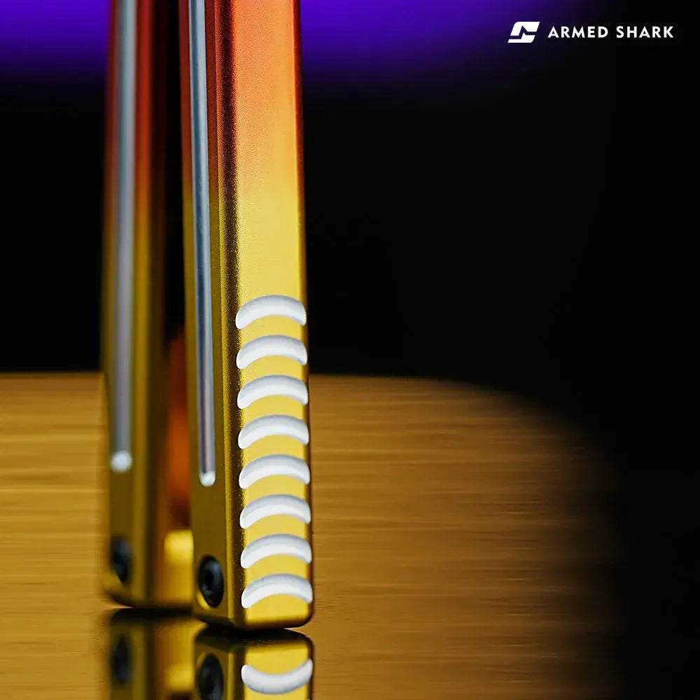 Close-up of Armed Shark Inked Mako balisong trainer handle tail with ladder jimping and gold–orange anodized gradient.