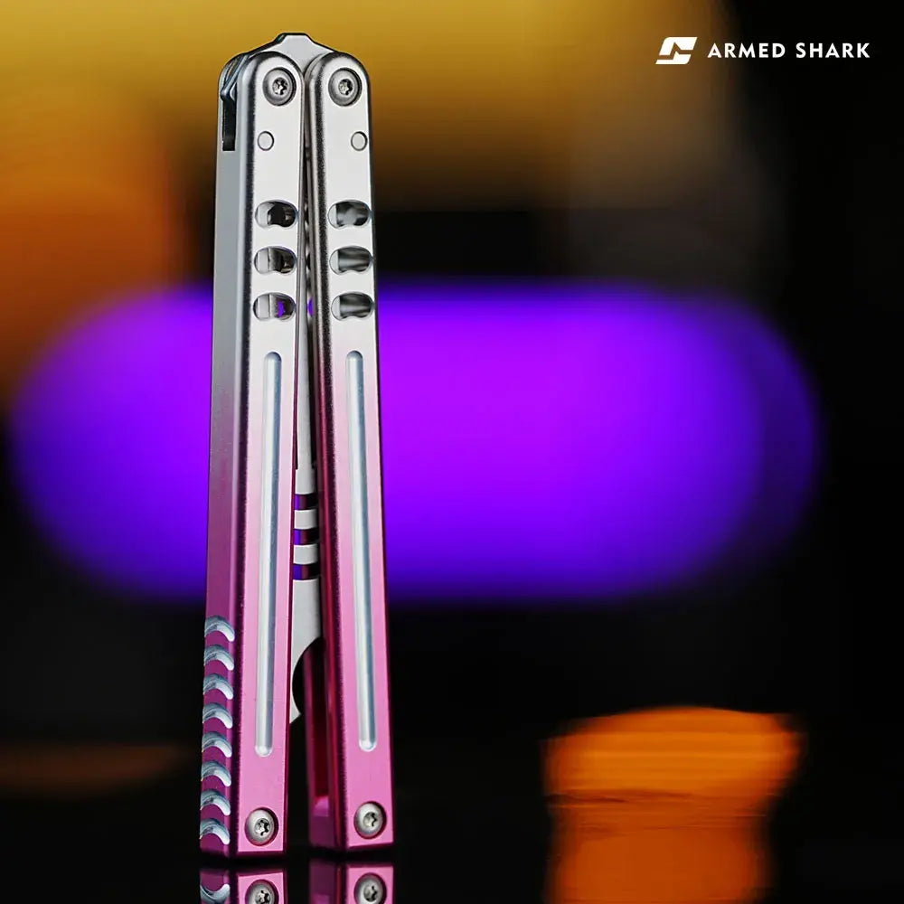 Armed Shark Inked Mako balisong trainer in silver-pink gradient, closed position, handle detail with slot milling, studio bokeh background.