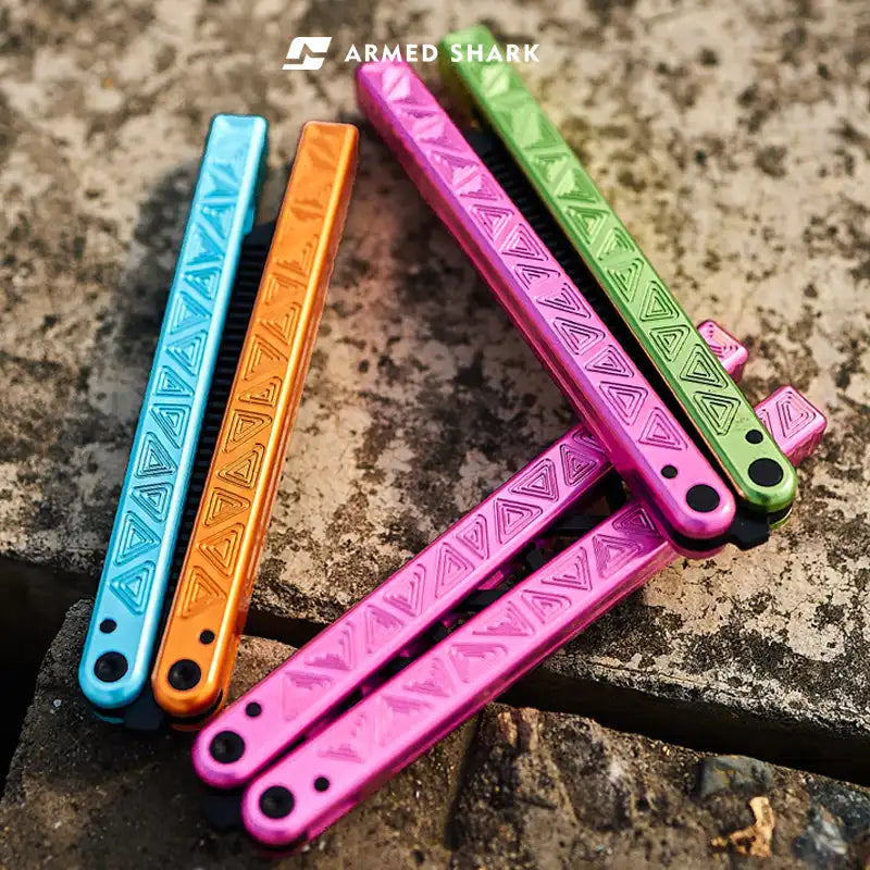 Colorful Armed Shark Original Balisong Trainer handles in pink, green, orange, and blue on outdoor stone surface.