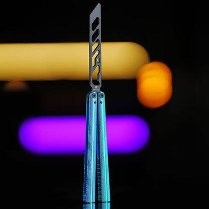 Armed Shark Pacific Ocean Balisong Trainer with blue anodized aluminum handles and black training edge standing upright on reflective black surface with vibrant yellow and purple background lights
