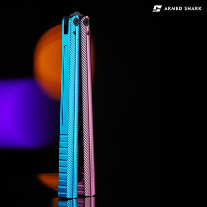 Back view of Armed Shark Pacific Ocean Balisong Trainer showcasing dual-tone anodized blue and pink aluminum handles with textured grooves, captured under warm orange and purple studio lighting for a premium metallic finish