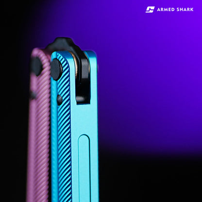 Macro close-up of Armed Shark Pacific Ocean Balisong Trainer pivot system showing dual-tone pink and blue anodized handles with precision bushing structure under violet studio lighting