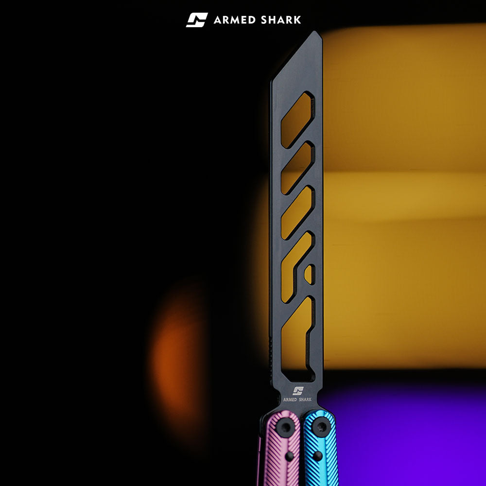 Close-up of Armed Shark Pacific Ocean Balisong Trainer showing pink-blue gradient anodized handles and black stainless training edge with precision cutouts, captured under warm orange and violet studio lighting