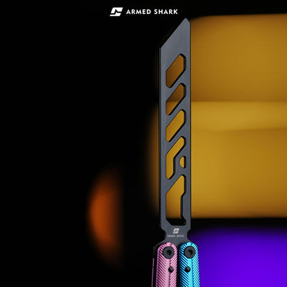 Close-up of Armed Shark Pacific Ocean Balisong Trainer showing pink-blue gradient anodized handles and black stainless training edge with precision cutouts, captured under warm orange and violet studio lighting