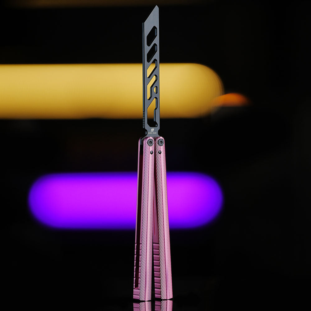 Armed Shark Pacific Ocean Balisong Trainer with pink anodized aluminum handles and black training edge standing upright on reflective surface with yellow and purple background lights