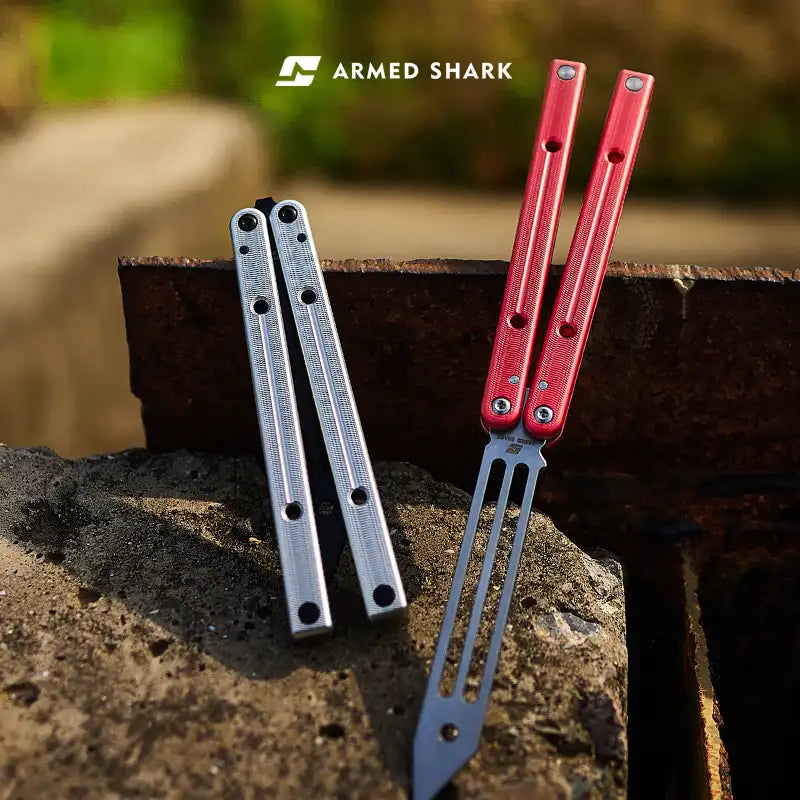 Outdoor shot of Armed Shark Squidtrainer V4 7075 aluminum balisong trainers: red unit open with slotted training head and silver unit closed on a concrete ledge.