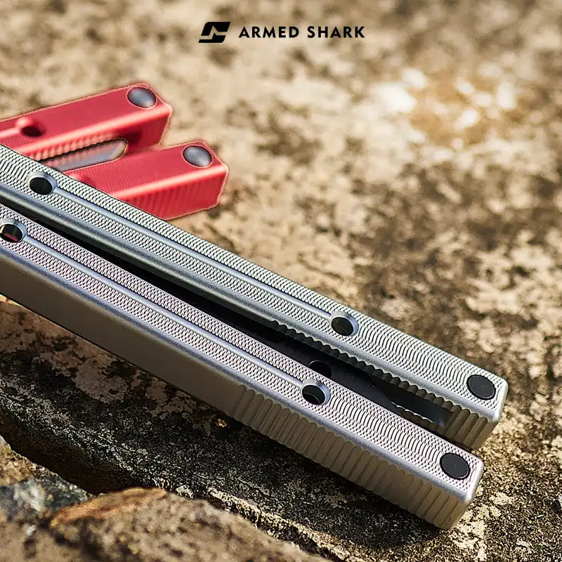 Close-up of Armed Shark 7075 aluminum balisong trainer handles in silver, with red handle in the background on a stone surface.